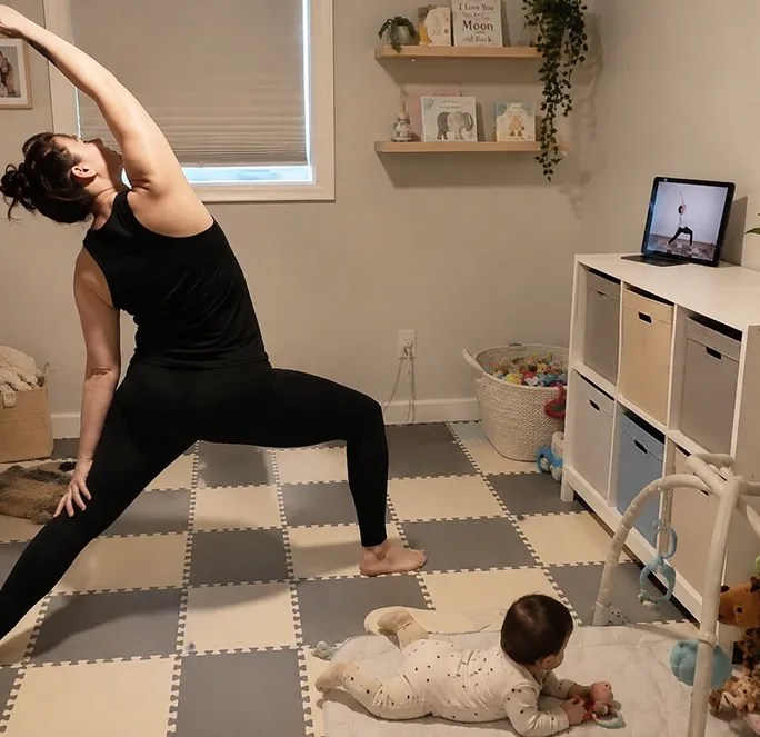 Parent practicing an online Power Yoga Collective class at home while caring for a baby in a nursery, demonstrating how on demand yoga fits into everyday family life.